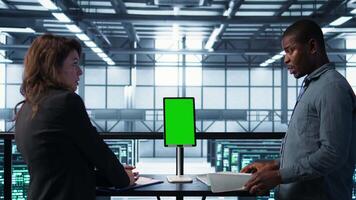 Programmers in data center brainstorming, using mockup tablet, observing equipment analytics. Server hub IT coworkers use isolated screen device, adjusting system parameters, camera B video