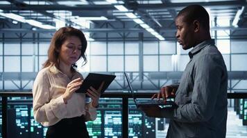 Smiling admins using laptop in data center, managing server virtualization. Cheerful IT specialist and african american man using notebook in server room, ensuring infrastructure reliability, camera B video