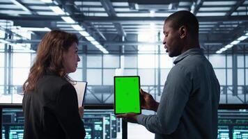 Server room data scientists using mockup tablet, conducting security audits to prevent bugs. African american man with colleague in data center looking at green screen device, camera B video