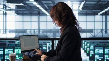 Admin in data center using laptop to update storage arrays, configuring network virtualization. Woman in server room holding coffee cup, verifying settings to maintain stable performance, camera B video