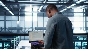 Engineer in data center with laptop automating routine maintenance tasks, executing diagnostic scripts. Man coding on notebook, adjusting power settings, validating server configurations, camera B video