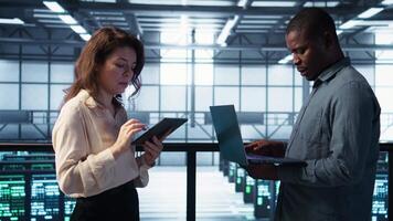 Portrait of happy system administrators in data center using devices, verifying firewall configurations. Jolly teamworking IT experts performing equipment vulnerability scans in server hub, camera B video