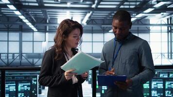 Data center management executives reading paperwork files, reviewing system diagnostics. Server farm colleagues looking over maintenance schedules documents and cybersecurity protocols, camera B video