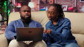 African american employees in living room using laptop and preparing presentation, inserting charts and adjusting layouts. Young couple finishing company project and writing summaries. Camera B. video