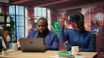 African American woman explores virtual reality using a VR headset while her partner works remotely on a laptop. Young couple embraces modern technology and gadgets with AI at home. Camera A. video