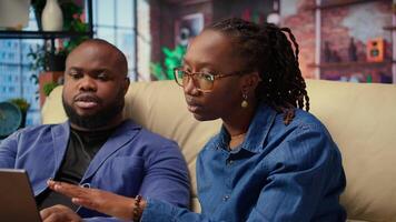 African american employees in living room using laptop and preparing presentation, inserting charts and adjusting layouts. Young couple finishing company project and writing summaries. Camera A. video