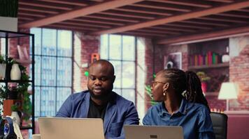 Black man and woman sits together in their cozy apartment space, discussing digital marketing strategy on a laptop. Supporting each other in their remote work and freelancing journey. Camera B. video