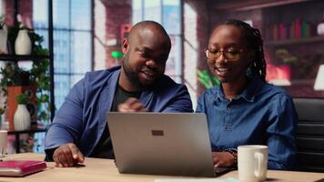 Black man and woman working in their cozy loft discussing digital marketing strategy on a laptop, pointing at online tools. Supporting each other in their remote freelancing journey. Camera B. video