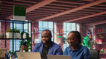 Black man and woman sits together in their cozy apartment space, discussing digital marketing strategy on a laptop. Supporting each other in their remote work and freelancing journey. Camera A. video