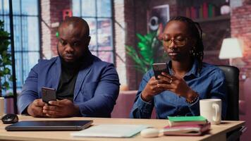 Young adults in their living space browsing webpages on smartphones together while they sit at the desk, having fun and enjoying leisure time with their gadgets. Cozy apartment. Camera B. video