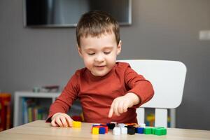 A cute happy little toddler boy of two years old sits at a children's table and plays with multi-colored blocks in his room. Educational toys for children. Soft selective focus photo