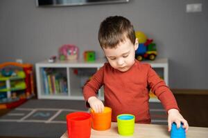 A cute happy little toddler boy of two years old sits at a children's table and plays with multi-colored pyramids in his room. Soft selective focus photo