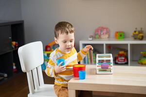 A cute happy little toddler boy of two years old sits at a children's table and plays with multi-colored pyramids in his room. Educational toys for children photo