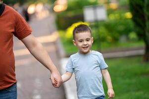 A happy three and a half years son walks hand in hand with his dad in the park on a sunny summer day. Family fun time and leisure. Summertime. photo