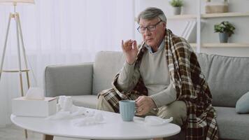 An elderly man in his 60s sits on a sofa wrapped in a blanket, looking tired and unwell. He holds his head, surrounded by tissues and medicine, in a bright living room. video