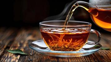 A steaming cup of tea being poured from a glass teapot on a rustic wooden table photo