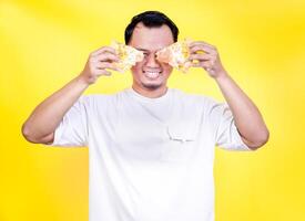 A cheerful asian man playfully holds pizza slices in front of his face against a vibrant yellow backdrop. international foods day concept photo