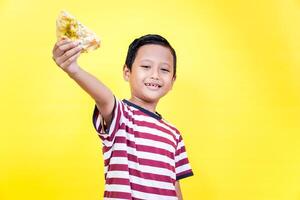 A cheerful asian boy holds up a slice of pizza against a bright yellow background, spreading joy. international foods day concept photo
