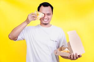 An asian man joyfully holding a slice of pizza in front of his face against a bright yellow background. international foods day concept photo
