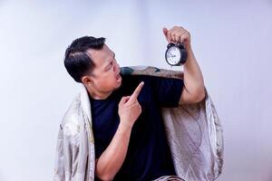 An asian man, surprised and expressive, holds an alarm clock while wrapped in a blanket isolated on white photo