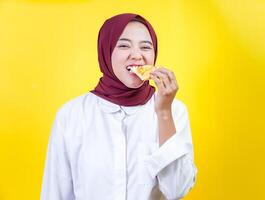 A joyful asian woman in a hijab enjoys a slice of pizza against a bright yellow background photo