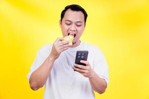 An asian man enjoys a slice of pizza while looking at his smartphone against a yellow background photo