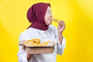 An asian woman bites a slice of pizza while holding a box of pizza in front of a bright yellow background. photo