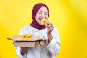 asian woman enjoys a slice of pizza while holding a box of pizza in front of a bright yellow background. photo