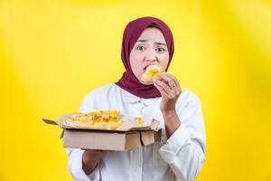 An asian woman in a hijab enjoys a slice of pizza while holding a box of pizza in front of a bright yellow background. photo