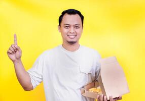 A smiling asian man in a white shirt holds a pizza box, pointing up against a vibrant yellow background. international foods day concept photo
