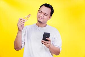 a man holding a piece of pizza and looking at his phone photo