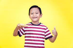 A cheerful asian boy in a striped shirt holds a slice of pizza and gives a thumbs up photo