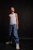 A young Asian boy stands in a studio against a black background, wearing jeans and a white tank top. His confident expression and relaxed posture create a striking portrait. photo