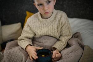 Wrapped in a blanket, the boy sits in bed, enjoying the soothing warmth of his tea with lemon. photo