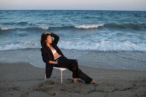 An elegant Asian woman in a strict suit stands against the backdrop of the ocean at sunset, her expression calm and powerful. photo