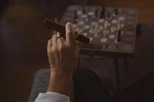 A steady hand holds a smoldering cigar, wisps of smoke curling above the chessboard, where a game of strategy unfolds in the dim light. photo