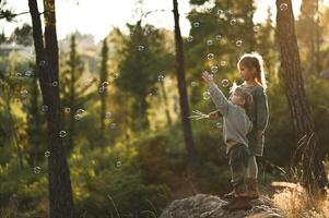 Among towering trees and soft moss, a boy and girl sit quietly, watching as shimmering bubbles drift like tiny fairy lights around them. photo