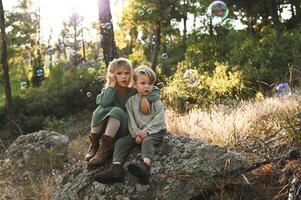 The boy and girl sit close together on an ancient stone, mesmerized by the rainbow-colored bubbles dancing around them in the enchanted forest. photo