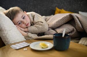 Lying in bed, the boy looks weak and tired. A mug of steaming tea with lemon and a few tablets sit on the table beside him. photo