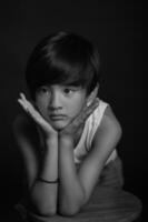 An Asian boy in a white tank top and jeans stands in a studio against a black background, his posture relaxed and natural, capturing a timeless portrait photo