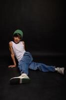 The boy, dressed in casual jeans and a white tank top, poses in a studio setting with a dark backdrop. His gaze is calm and focused. photo