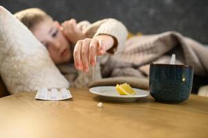 The sick boy lies in bed under a warm blanket. In front of him, on the bedside table, there is a cup of hot tea with lemon and some medicine. photo