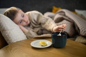 The boy rests in bed, wrapped in a blanket. On the bedside table, a warm cup of tea with lemon and medicine are placed, helping him recover photo