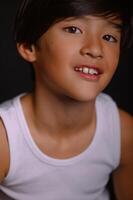 With a black background behind him, a boy of Asian descent poses in a white tank top and jeans, his expression both thoughtful and natural. photo