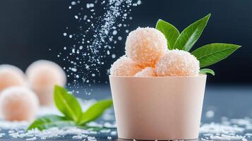 Close-up of coconut covered dessert balls in a paper cup with fresh green leaves, surrounded by falling sugar and a blurred dark background. photo