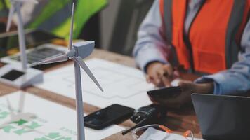 A man in an orange vest is working on a laptop and a wind turbine video