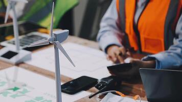 A man in an orange vest is working on a laptop and a wind turbine video