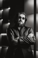 Portrait of handsome, young, confident bearded man adjusting in glasses, looking at camera while standing against dark studio background. Black and White photo