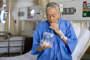 Senior asian man using the incentive spirometer device for breathing exercise rehabilitation to recover from lung cancer after surgery due to heavy smoking in hospital bed photo