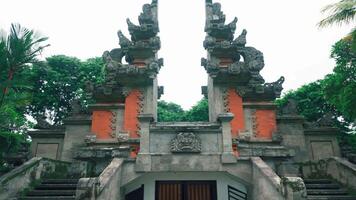 A traditional Balinese temple entrance with intricate stone carvings and vibrant greenery surrounding it. The structure features ornate details and a symmetrical design video